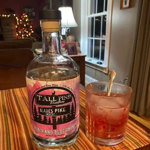 A bottle of Tall Pines Distillery liquor next to a glass of mixed drink on a table, with a festive background.