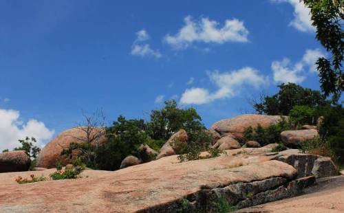 A rocky landscape under a bright blue sky with scattered clouds and greenery in the background.