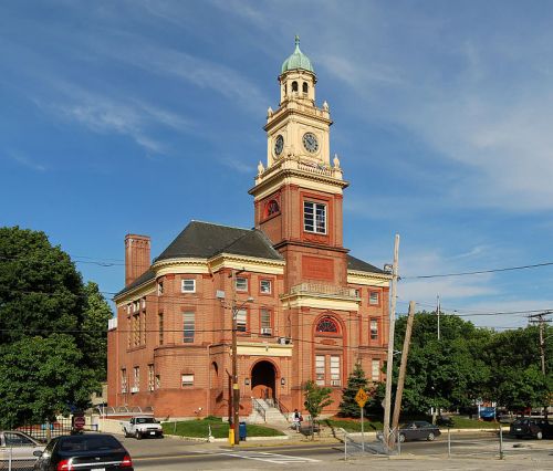 Historic red brick building with a clock tower, surrounded by trees and cars under a clear blue sky.