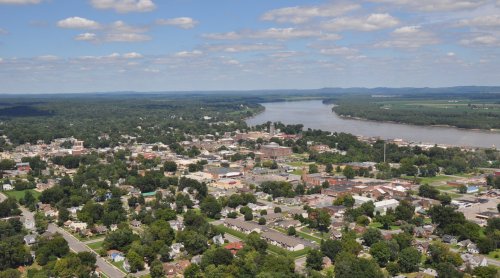 Aerial view of a small town by a river, surrounded by greenery and rolling hills under a partly cloudy sky.