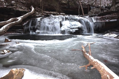 A frozen waterfall surrounded by icy rocks and a snow-covered landscape, with a fallen tree in the foreground.