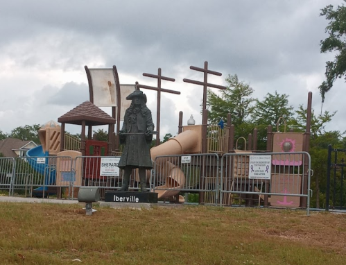A playground with colorful structures and a statue of a historical figure in front, surrounded by fencing.
