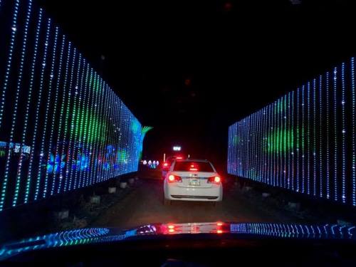 A car drives through a colorful light tunnel at night, with vibrant blue and green lights illuminating the surroundings.