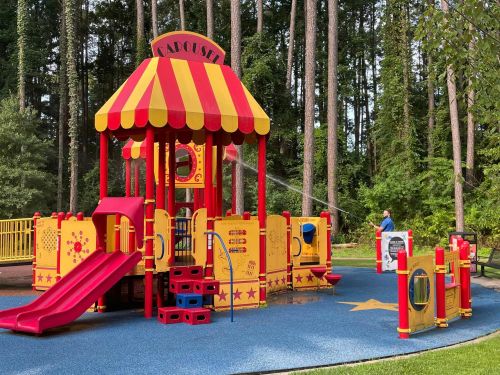 Colorful playground with a carousel structure, slides, and play areas, surrounded by trees in a park setting.