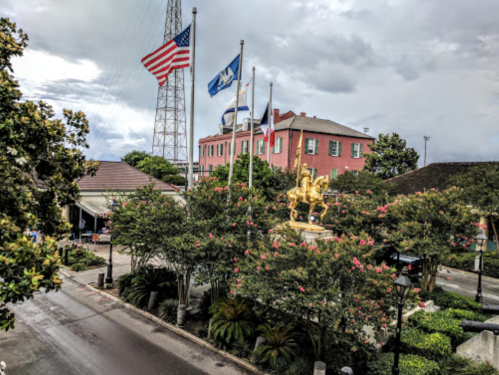 View of a park with trees, flags, and a golden statue, set against a cloudy sky and a historic building in the background.