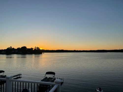 A serene lake at sunset, with calm waters reflecting the orange sky and silhouettes of trees in the distance.