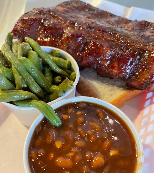 A plate featuring a juicy rib, green beans, baked beans, and a slice of bread.