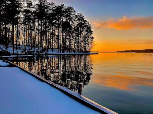 A serene lakeside scene at sunset, with snow-covered banks and trees reflecting on the calm water.