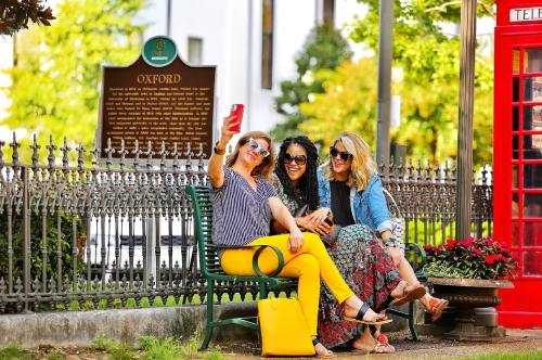 Three women take a selfie on a bench in a park, surrounded by greenery and a historic sign in the background.