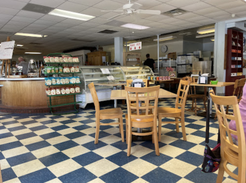 A cozy café interior with wooden tables, chairs, and a display case, featuring a checkered floor and a counter in the background.