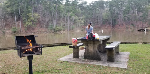 A person prepares food at a picnic table near a lake, with a grill smoking in the foreground and trees in the background.
