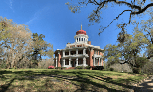 A grand, historic mansion with a red dome, surrounded by trees and blooming flowers under a clear blue sky.