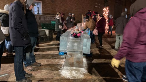 A crowd gathers around an ice sculpture table, with people enjoying a winter event in a festive atmosphere.