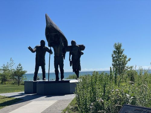 Three statues of figures holding a canoe, set against a clear blue sky and a lake in the background.