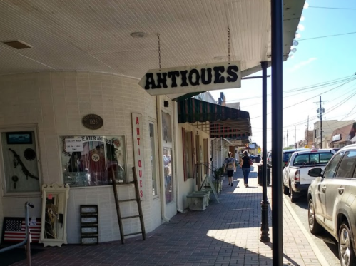 A street view of a quaint antique shop with a sign, surrounded by other shops and parked cars.