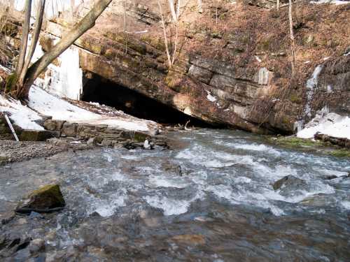 A flowing river with rocks and snow, surrounded by trees and a stone overhang in a wooded area.