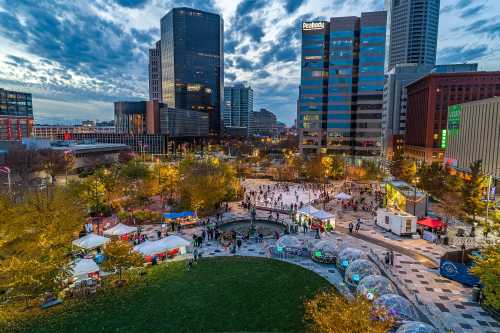 Aerial view of a vibrant city park with ice skaters, food stalls, and illuminated trees against a skyline at dusk.