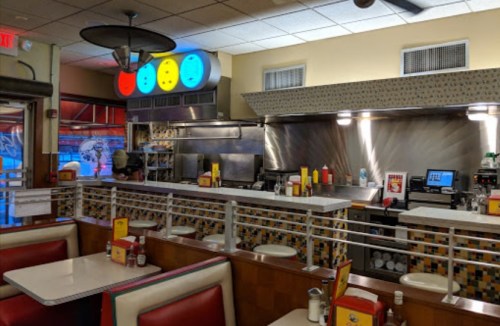A retro diner interior with red booths, a counter, and a kitchen area featuring colorful lights and condiments on tables.