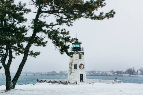 A snowy landscape featuring a lighthouse adorned with a wreath, surrounded by a calm body of water and a tree.