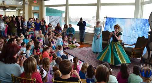 A crowd of children watches two performers dressed as princesses in a festive indoor setting.