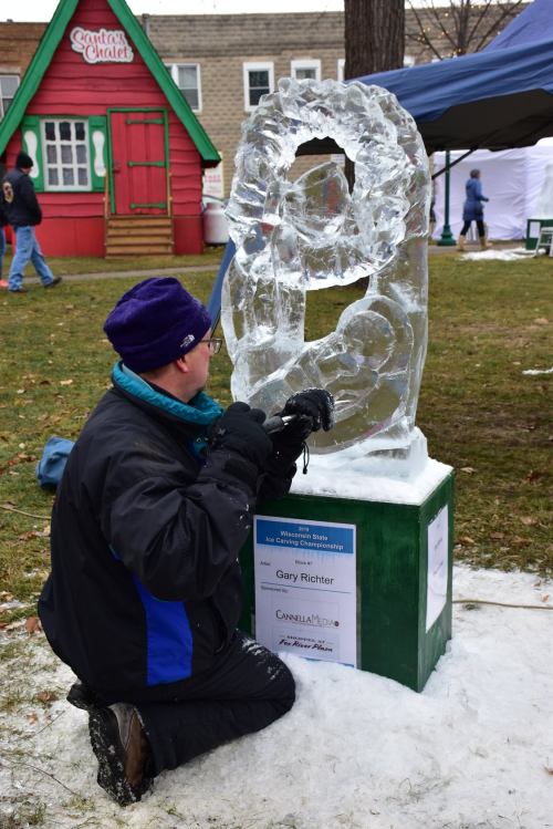 A sculptor carves an ice sculpture at an outdoor event, with a colorful chalet in the background.