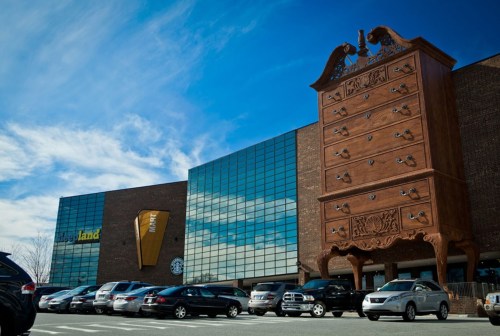 A large wooden dresser sculpture stands outside a modern building with reflective glass windows and parked cars.
