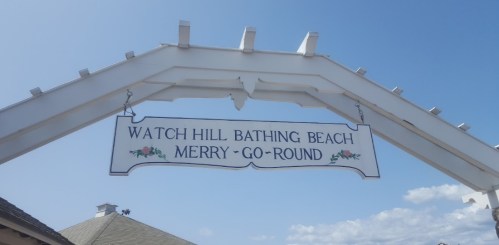 Sign for Watch Hill Bathing Beach and Merry-Go-Round under a blue sky with clouds.