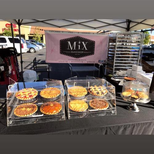 A display of various pies at a bakeshop booth, with a pink banner in the background and baked goods on a table.