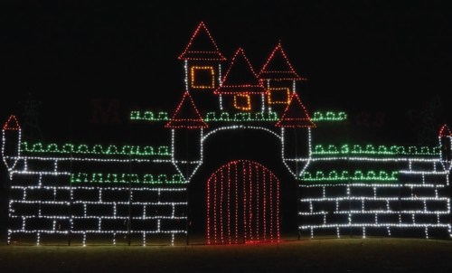 A brightly lit castle made of colorful holiday lights, featuring towers and a large gate against a dark night sky.