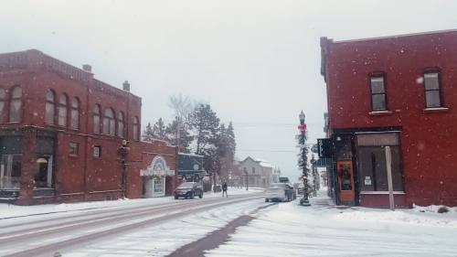Snowy street scene with red brick buildings, light snowfall, and a few pedestrians in a small town.