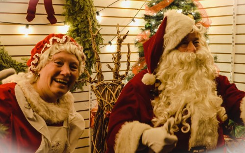 A cheerful Santa Claus and Mrs. Claus pose together in a festive setting with holiday decorations.