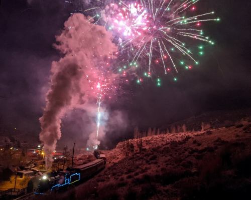 A train emits steam as colorful fireworks light up the night sky over a snowy landscape.