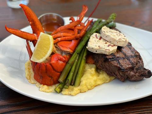 A gourmet plate featuring grilled steak, lobster, asparagus, and creamy grits, garnished with lemon and sauce.