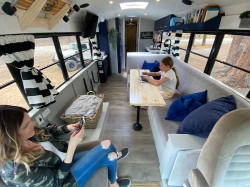 A cozy interior of a converted bus, featuring seating areas, a table, and two people using devices.