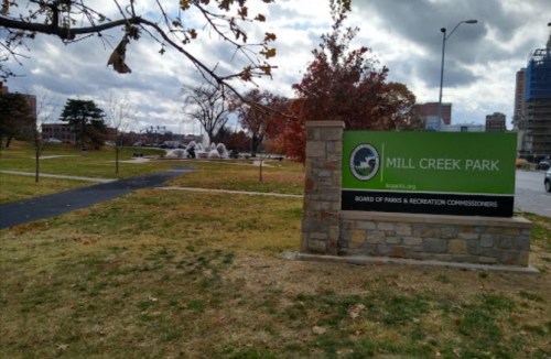 Sign for Mill Creek Park with a fountain in the background and a cloudy sky above. Trees are visible in the park.