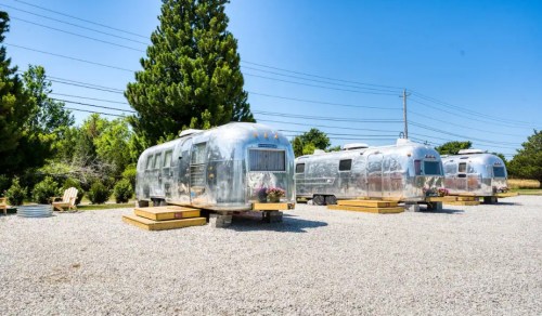 Three shiny Airstream trailers parked on a gravel lot, surrounded by greenery and under a clear blue sky.