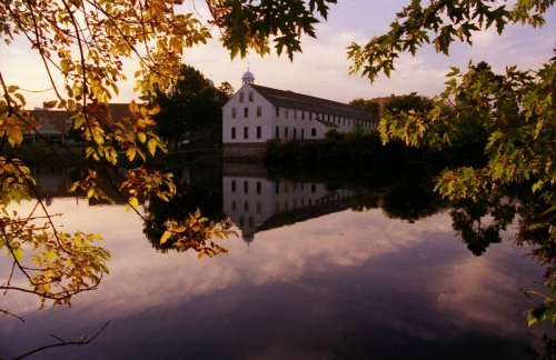 A serene scene of a white building reflected in calm water, framed by autumn leaves and a colorful sunset sky.