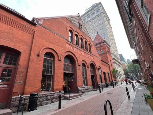 Historic red brick building with arched windows, labeled "Central Market," alongside a paved walkway and city skyline.