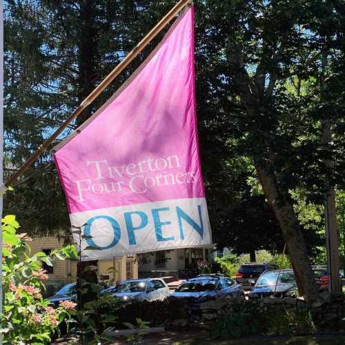 A pink flag with "Tiverton Four Corners OPEN" text, surrounded by greenery and parked cars in the background.