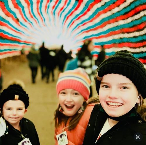 Three children smiling in front of a colorful, illuminated tunnel, wearing winter hats and jackets.