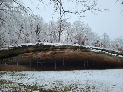 A snowy landscape featuring a large rock overhang and trees, with people visible on top of the hill.