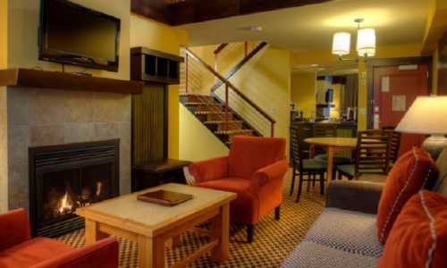 Cozy lounge area with a fireplace, orange chairs, wooden table, and a staircase in a warm, inviting setting.