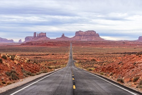 A long, straight road stretches through a desert landscape with red rock formations under a cloudy sky.