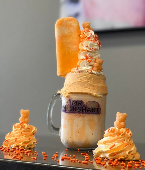 A tall milkshake topped with whipped cream, a popsicle, and orange sprinkles, accompanied by two decorative cupcakes.