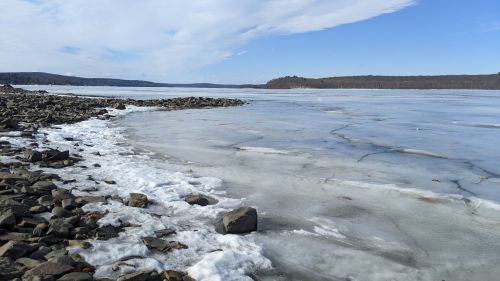 A frozen lake with rocky shores under a clear blue sky and distant hills. Ice and snow cover the water's surface.