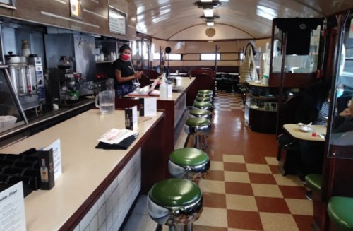 A retro diner interior with green stools, a counter, and a staff member wearing a mask. Patrons are seated in the background.