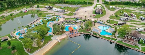 Aerial view of a resort with multiple pools, a lake, and RV parking surrounded by greenery.