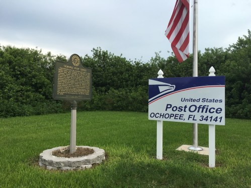 A historical marker and a sign for the Ochopee, FL Post Office, with a flag in the background and greenery.