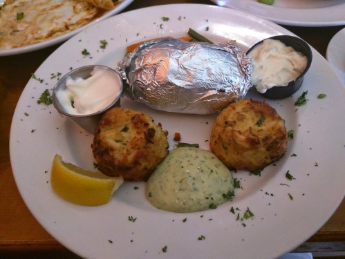 A plate featuring a baked potato, two crab cakes, lemon wedge, and sauces, garnished with herbs.