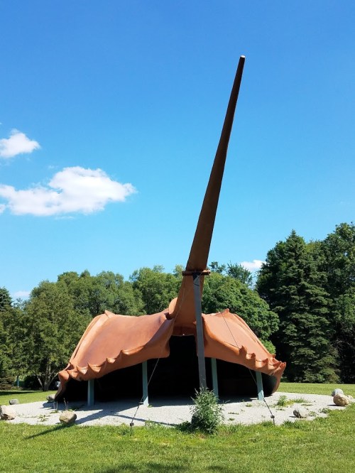 A large, brown, shell-like structure with a tall, pointed roof, set in a grassy area under a blue sky.